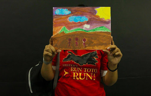 Child holding a painting that he created. His face is covered. It depicts him and his family waiting to cross the border.